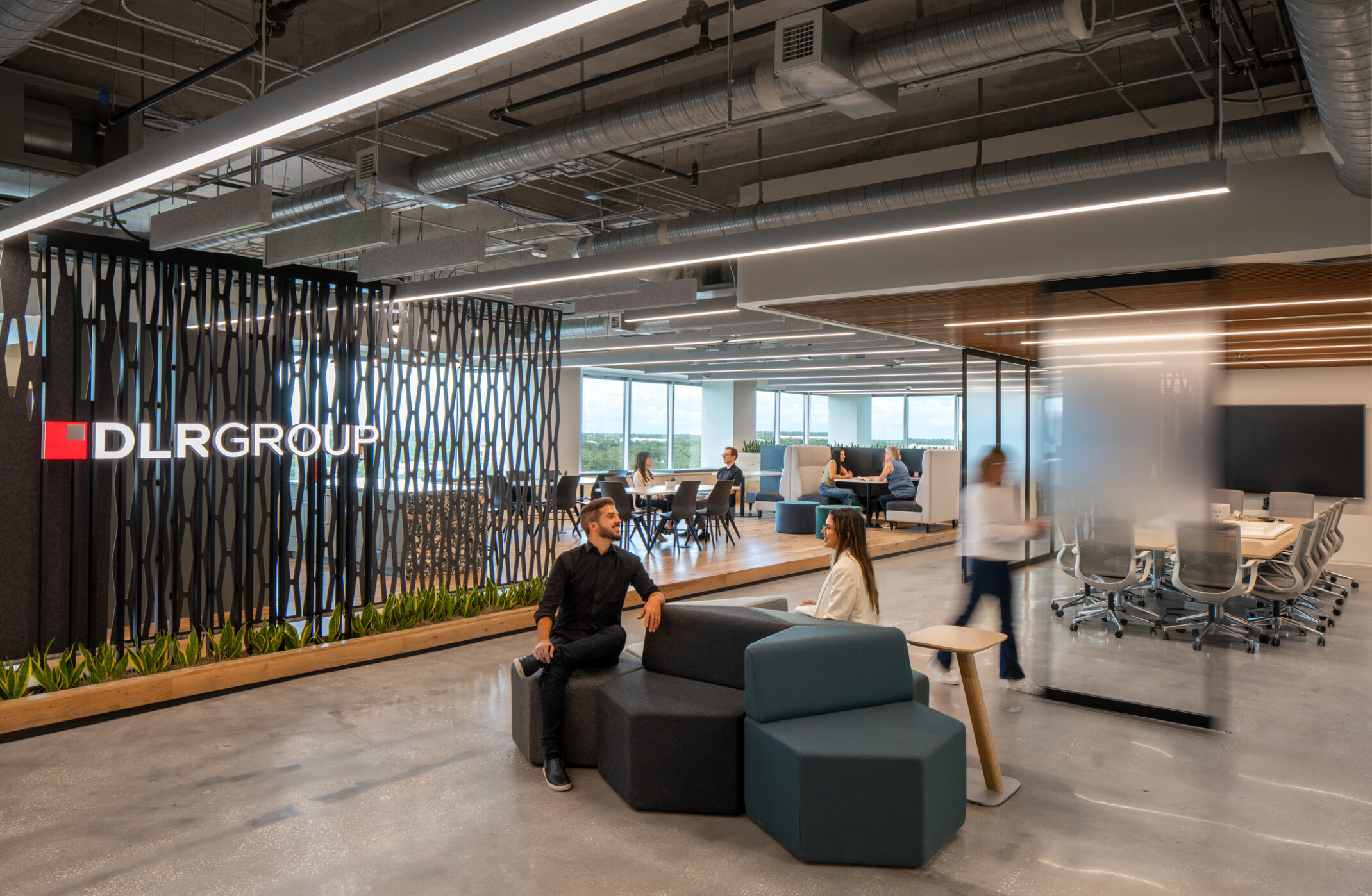 Modern office with modern artwork to the left with words DLR Group, dark low back couches in a seating area and people moving about