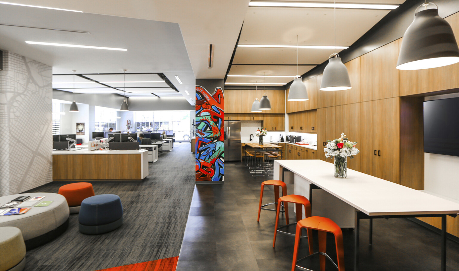 Office space with wood panel kitchen to right with counter height tables. Mural on center column, desks and seating to left Office space with wood panel kitchen to right with counter height tables. Mural on center column, desks and seating to left