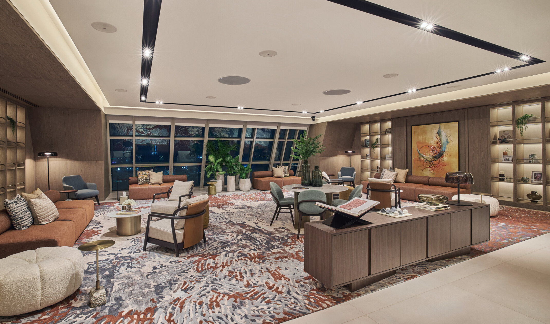 Hospitality-inspired office lounge with built-in shelving, colorful patterned rug, contemporary sofas, curated accessories, and floor-to-ceiling windows offering a city view.