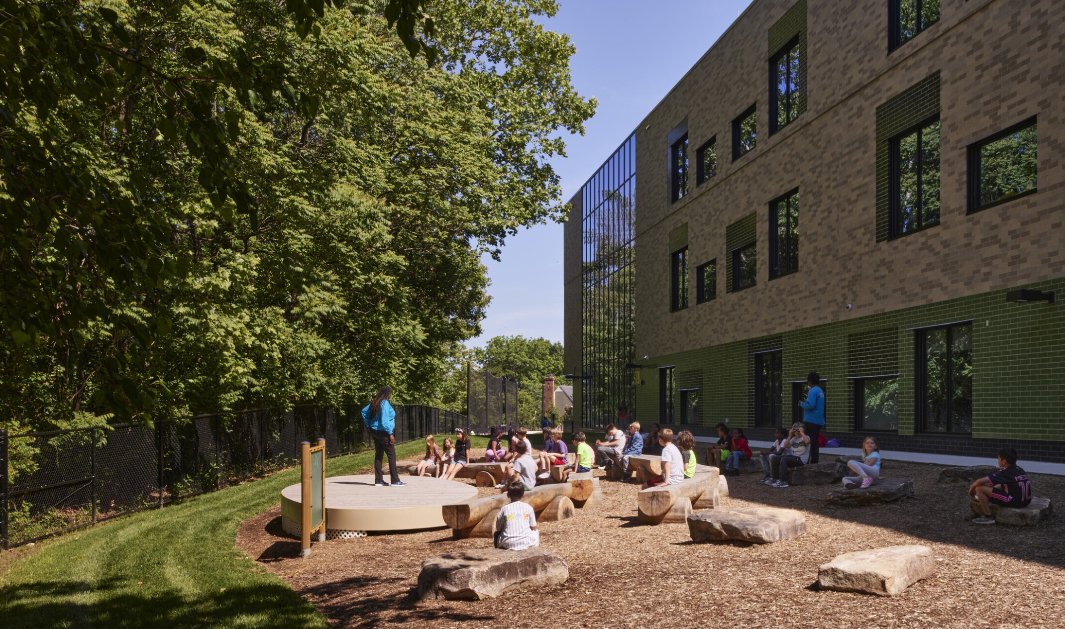 Outdoor learning area next to a multistory school filled with rocks and cut log seating looking at a densely wooded area