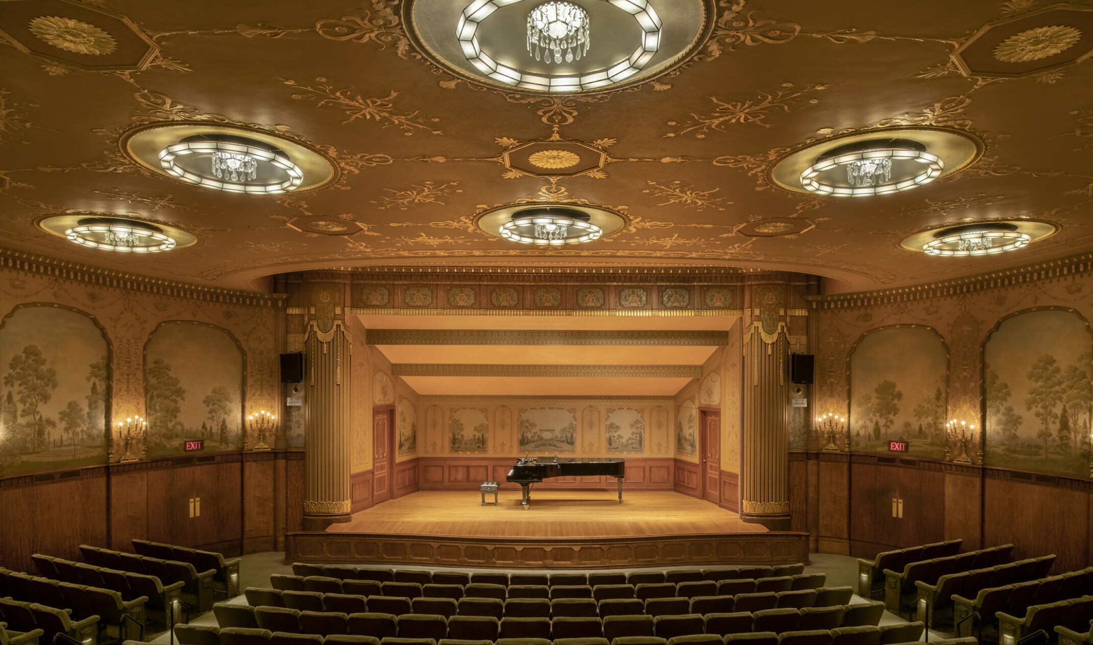 An intimate theater with a gold ceiling and crystal pendant lights. Tapestries and candelabra adorn walls and recital stage.