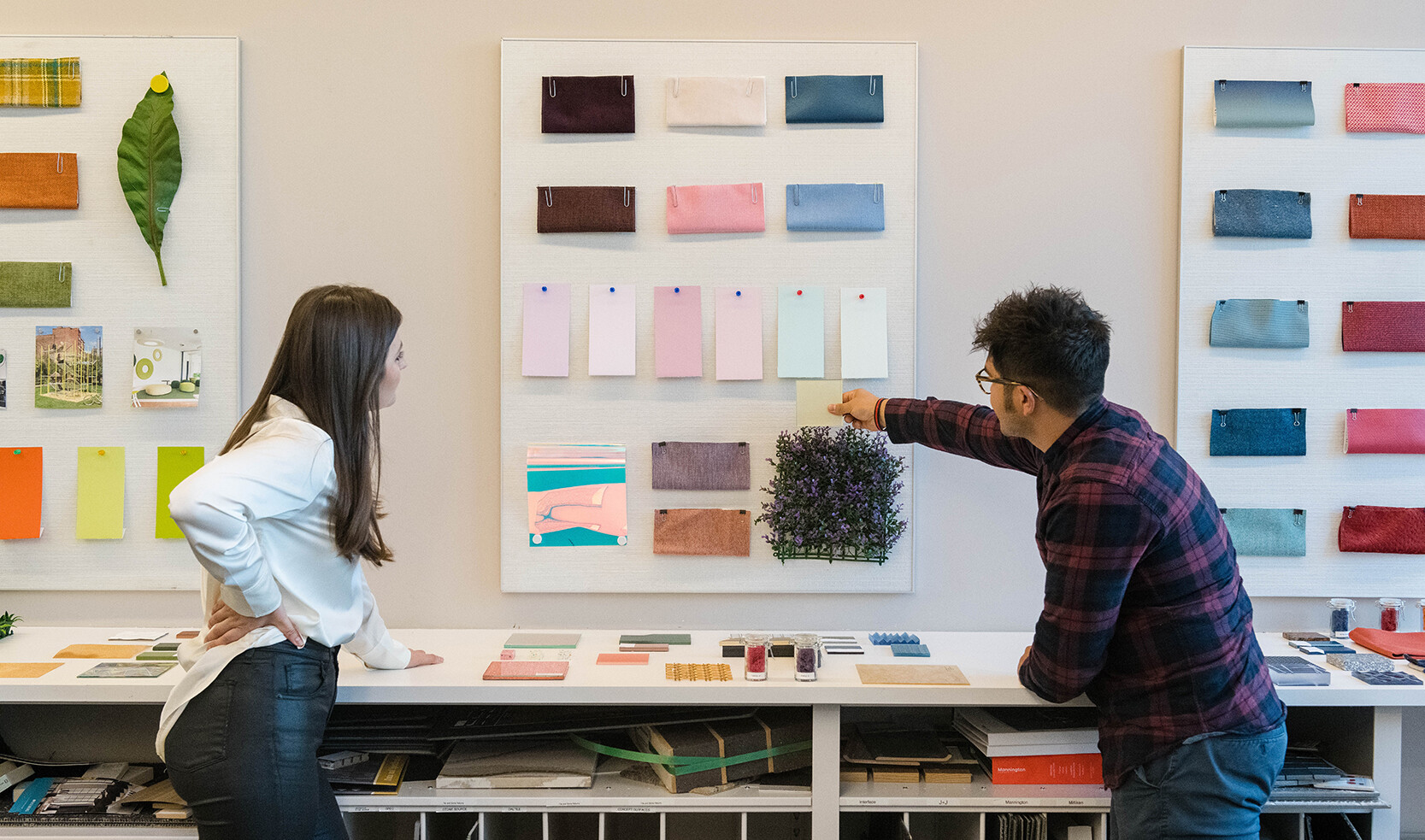 palette boards on walls with various color samples, inspiration images, and flowers and leaves pinned up. two people look on