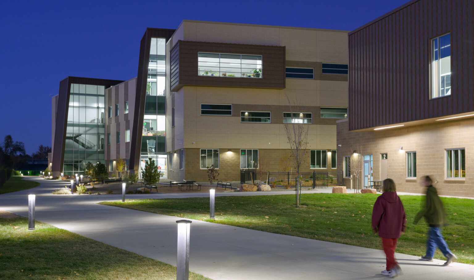 Grand Junction High School building at night; glowing light comes from inside walls of windows; two students walk toward the background buildings