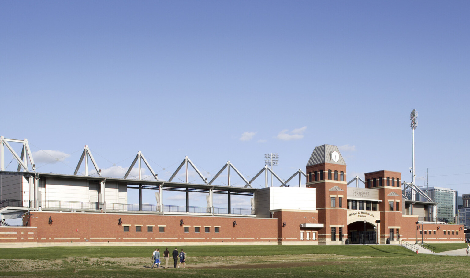 Arched stadium entry framed by brick towers and single story building. Behind, exposed 2nd floor walkway with vaulted beams