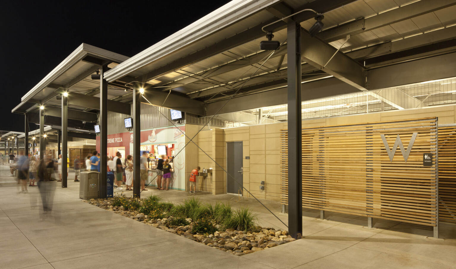 Silver awnings supported by exposed black beams over concession stands. Right, wood panel restroom entrance