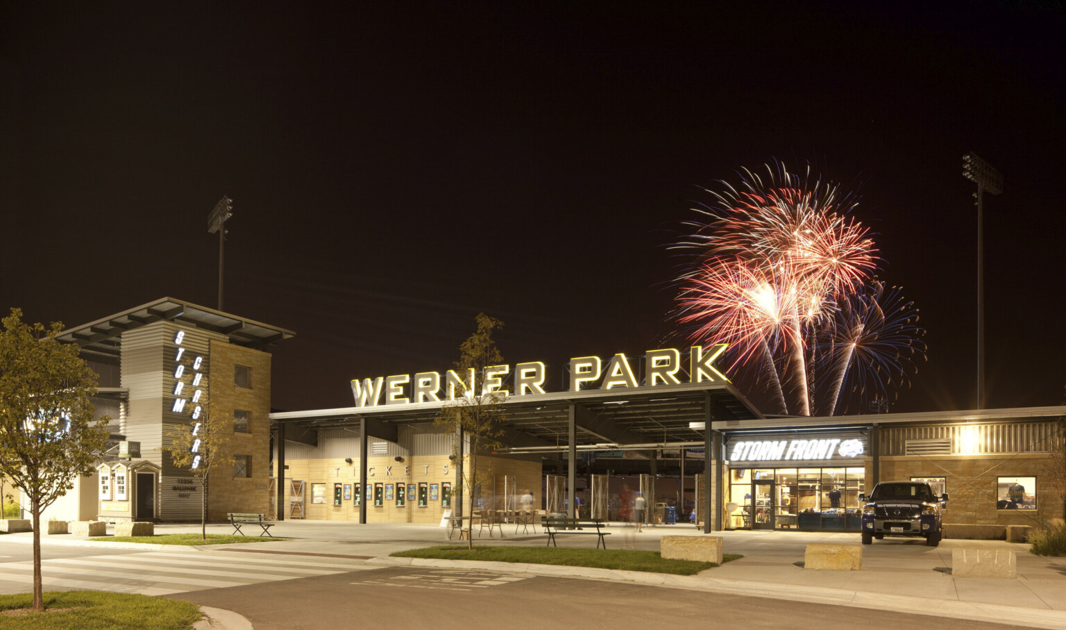 Stadium entrance with Werner Park sign illuminated at night. Right, Storm Front team store lit from within, fireworks above