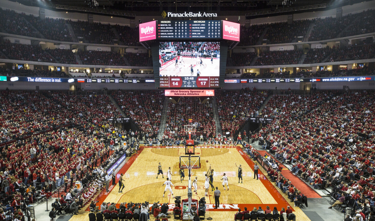 Basketball court during game with lighting and jumbotron hanging above. Bleachers surround the court and are filled with fans