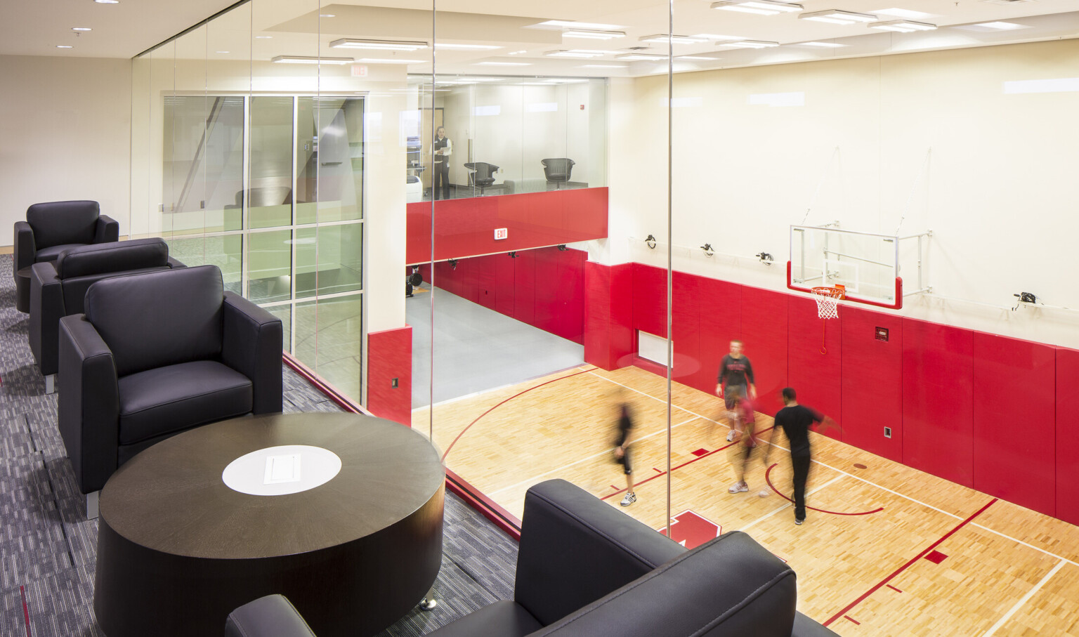 Black armchairs with tables line floor to ceiling windows of 2nd floor room looking down to indoor basketball half court