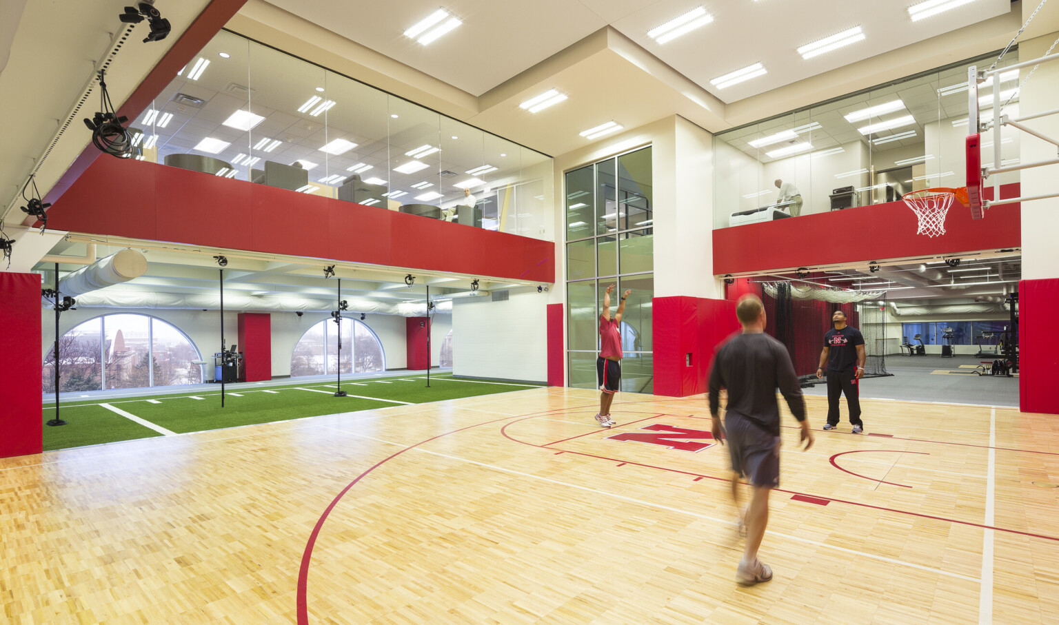 Double height basketball half court with windows on 2nd floor. Left, turf floor in front of arched windows with campus view
