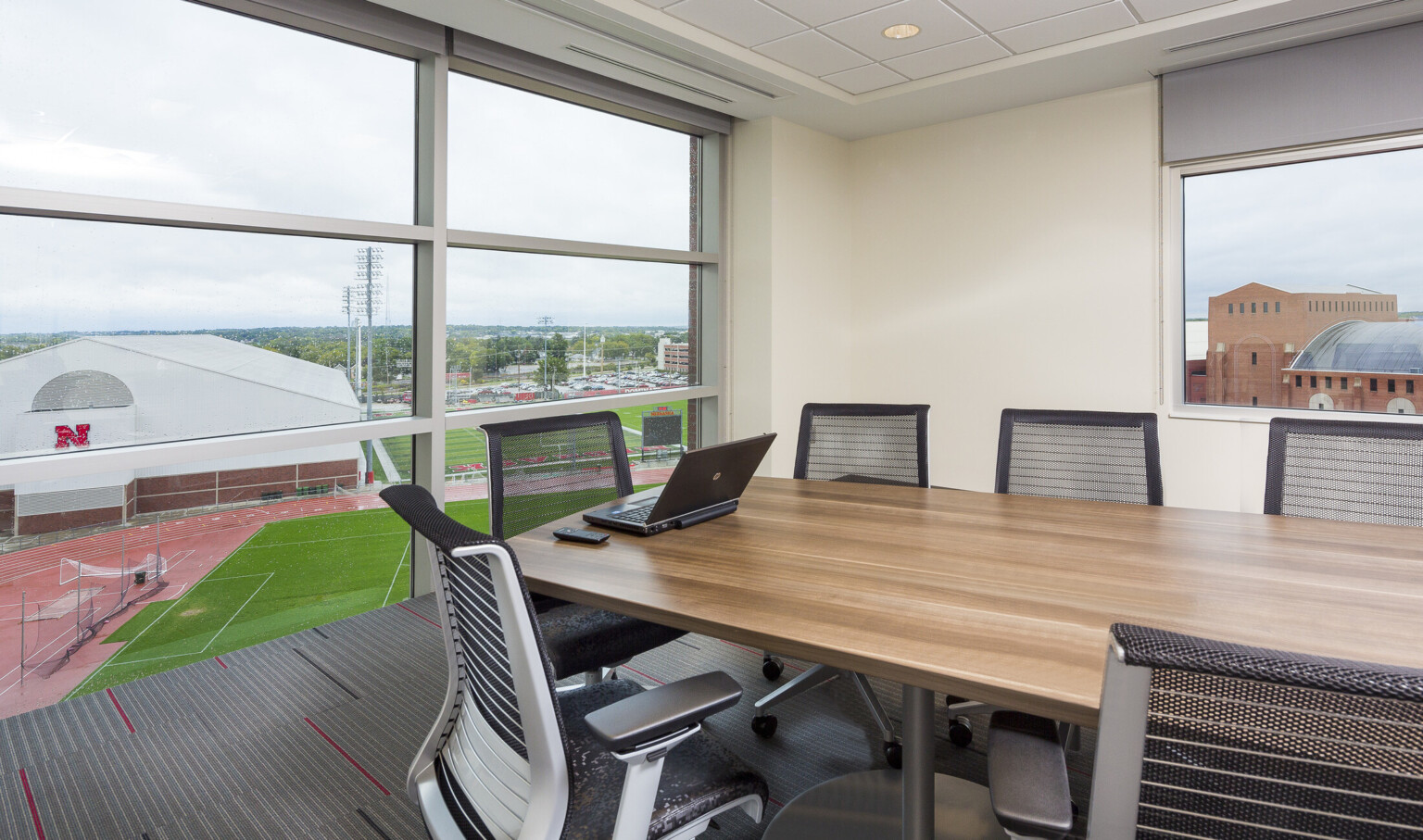 Corner conference room with wood table and black chairs. Floor to ceiling windows with white seams, left, view fields