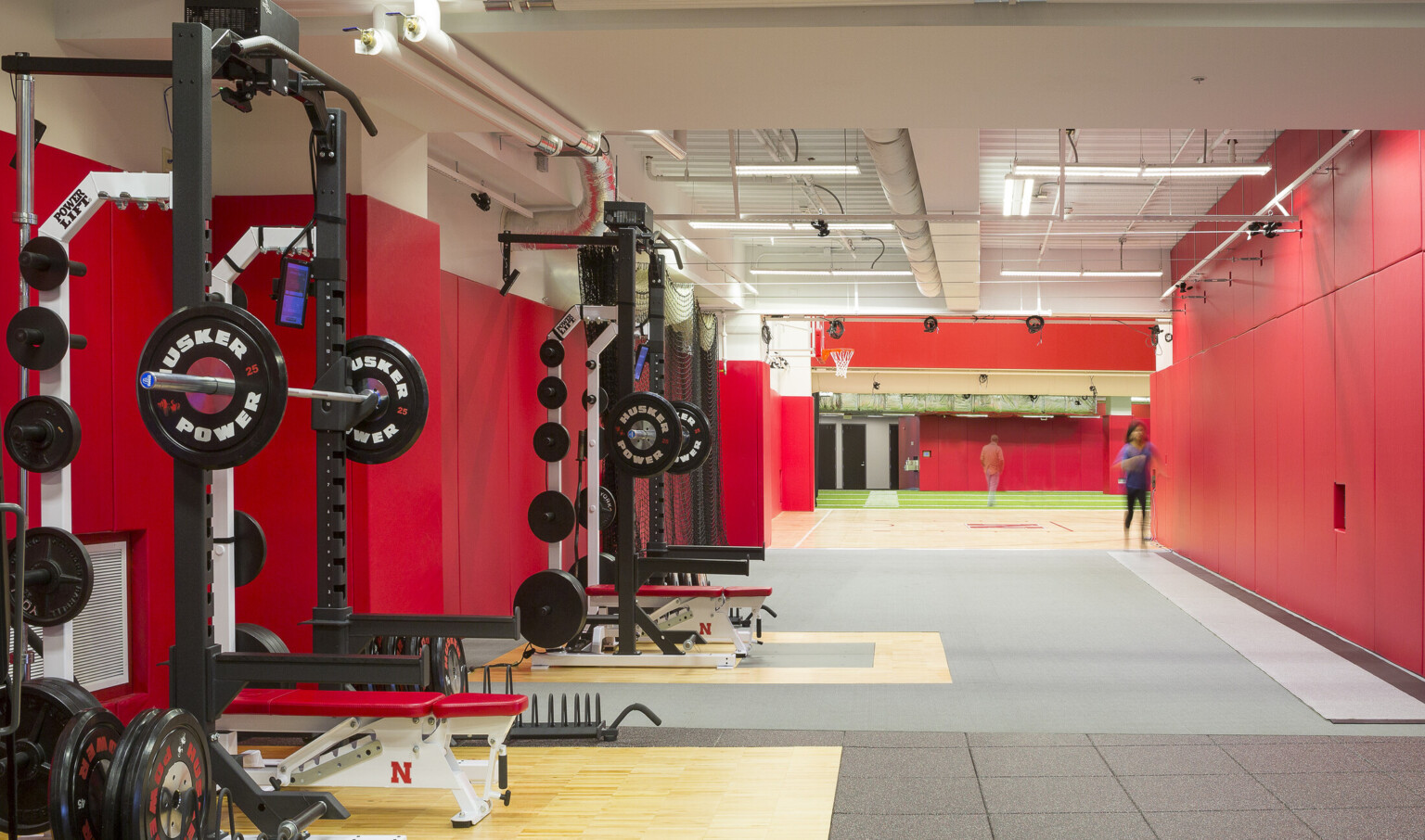 Exercise equipment in white room with red wall padding and exposed ductwork. Beyond, sections of hard wood and turf floor