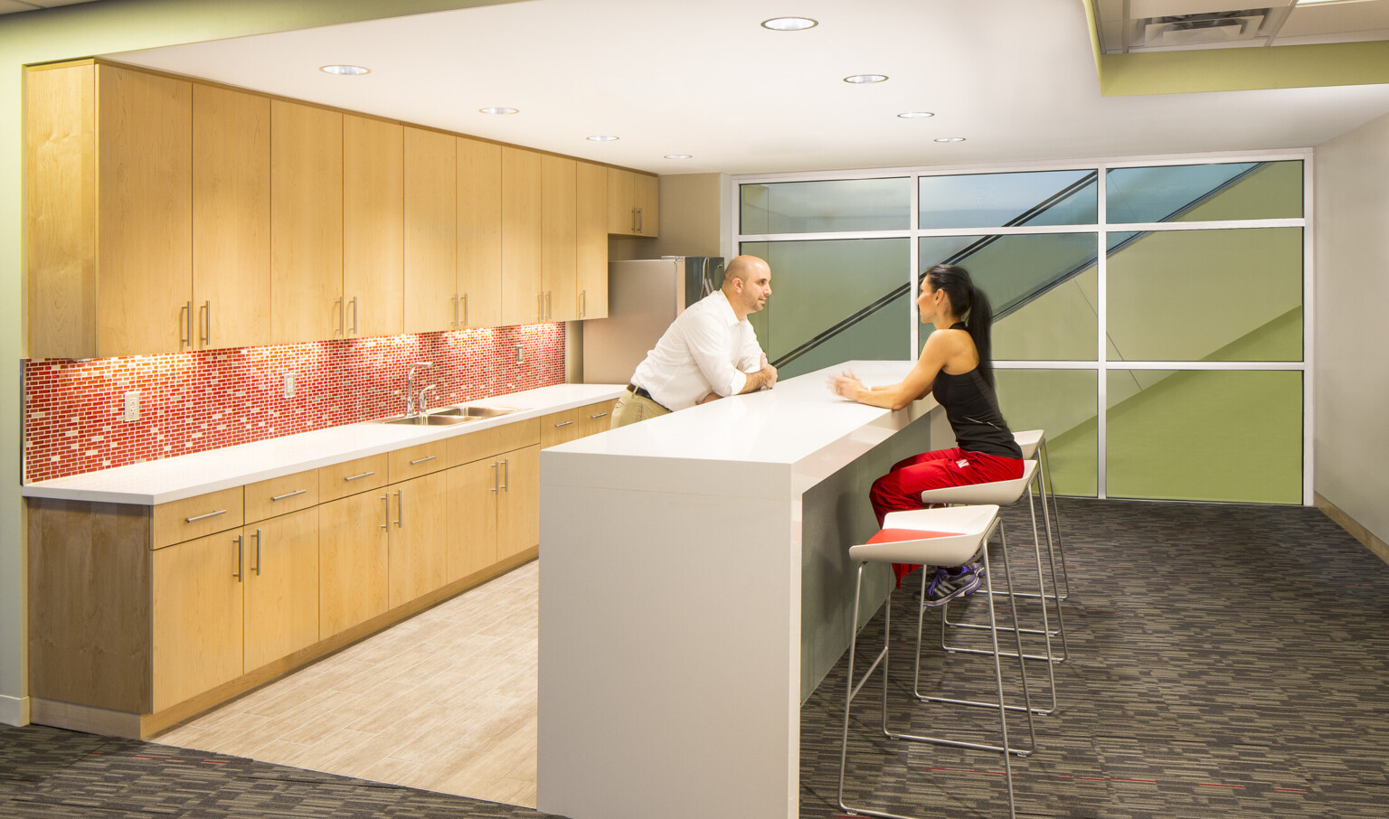 Kitchenette with red and white tiled backsplash between wood cabinets in white room. At center, island, with window, right