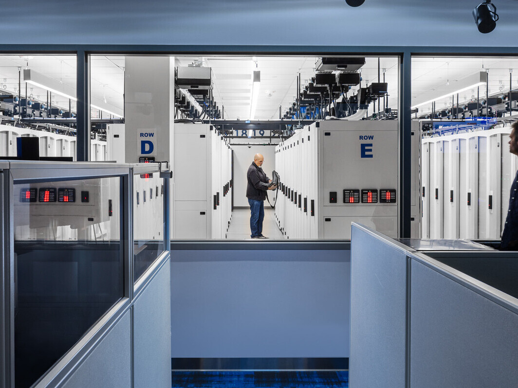 View of a data center interior with server racks and cubicles, featuring two individuals and labeled rows "D" and "E"
