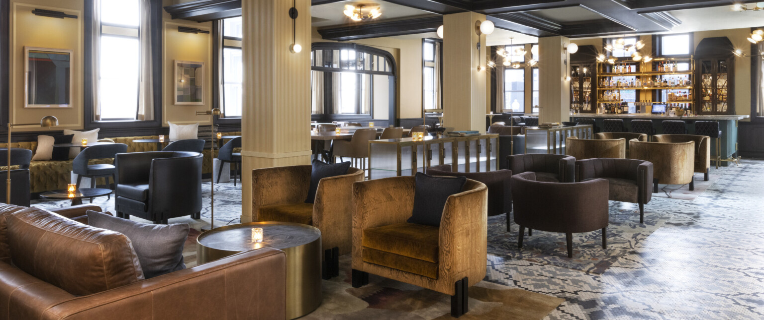 Seating area with arm chairs and round coffee tables and bar at back. Beige pillars below white ceiling with dark wood beams