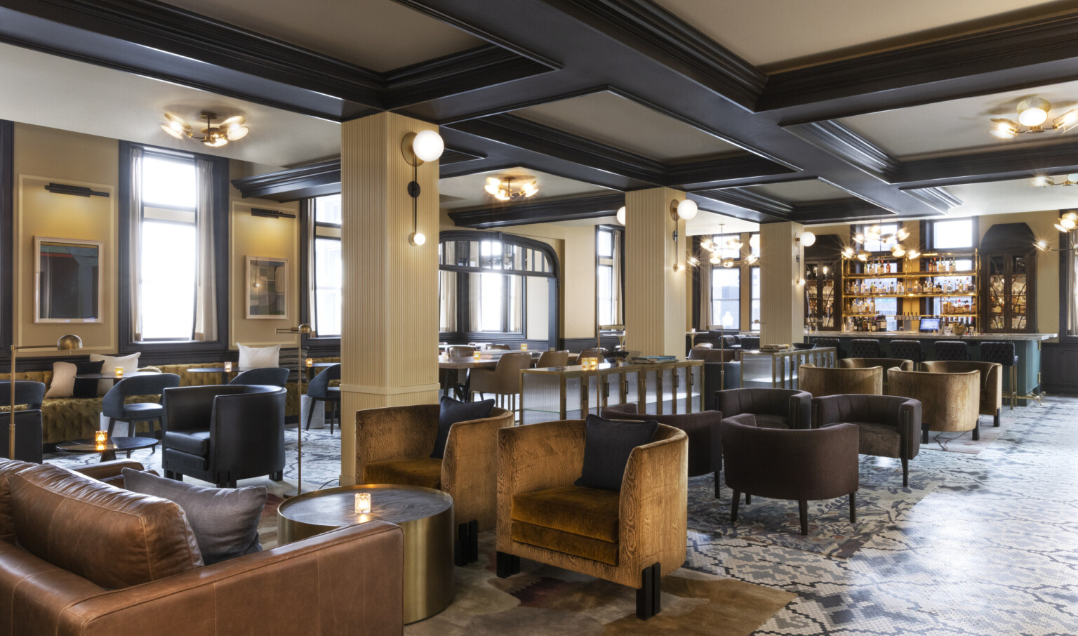 Seating area with arm chairs and round coffee tables and bar at back. Beige pillars below white ceiling with dark wood beams