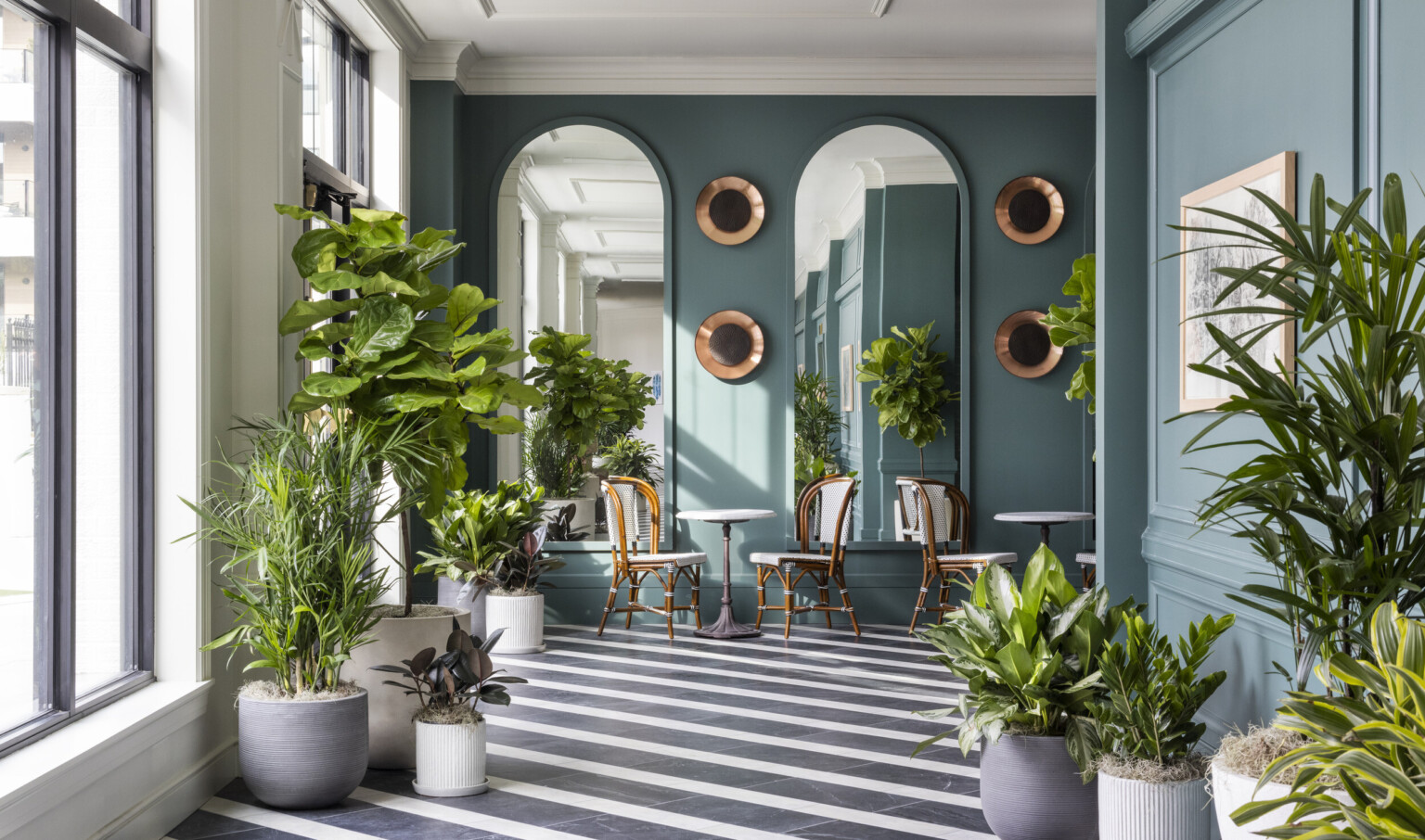 Black and white diagonal striped floor in green window lined hallway. Seating area and arched mirrors at end of hall