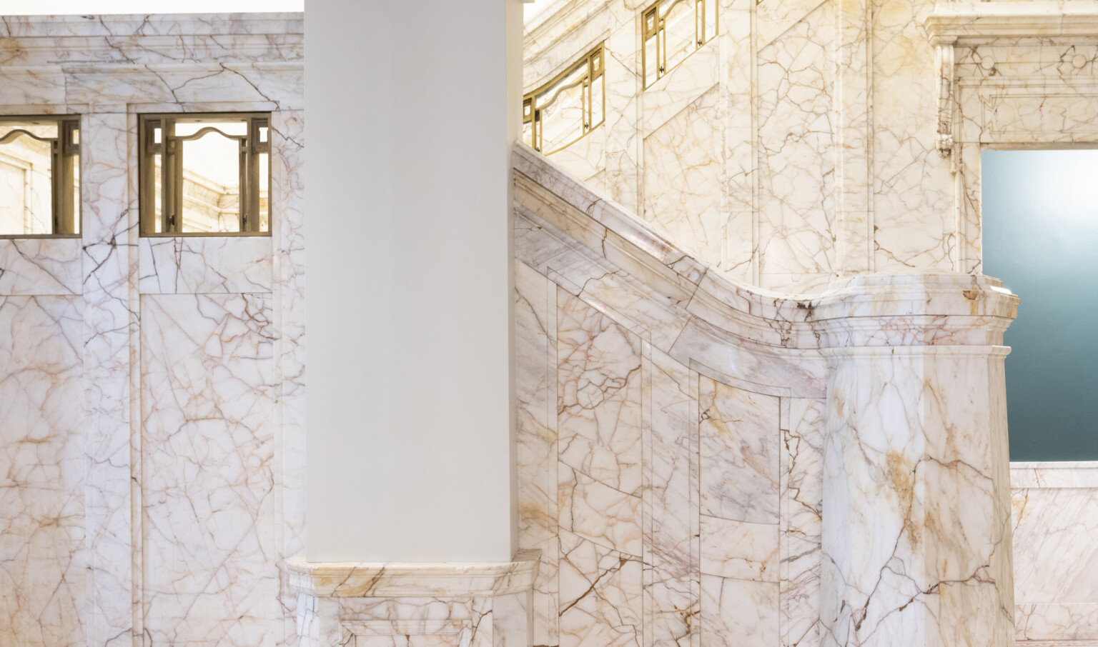 White column in a marble staircase with curving rail with sections of golden metal work inset
