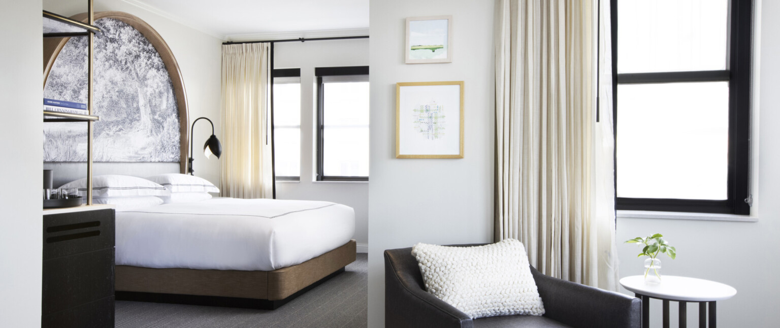Black arm chair with white knitted pillow in sitting area in front of bedroom with white bed on wood base and large windows