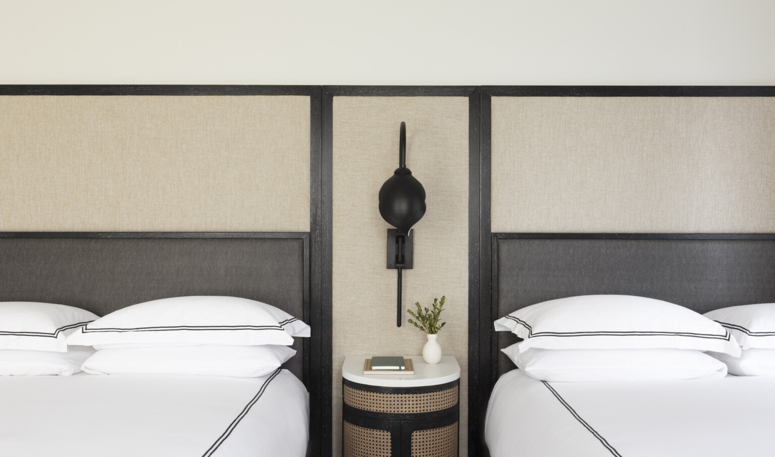 2 beds with white comforter and pillows with black double border against dark headboard and beige wall, separated by table