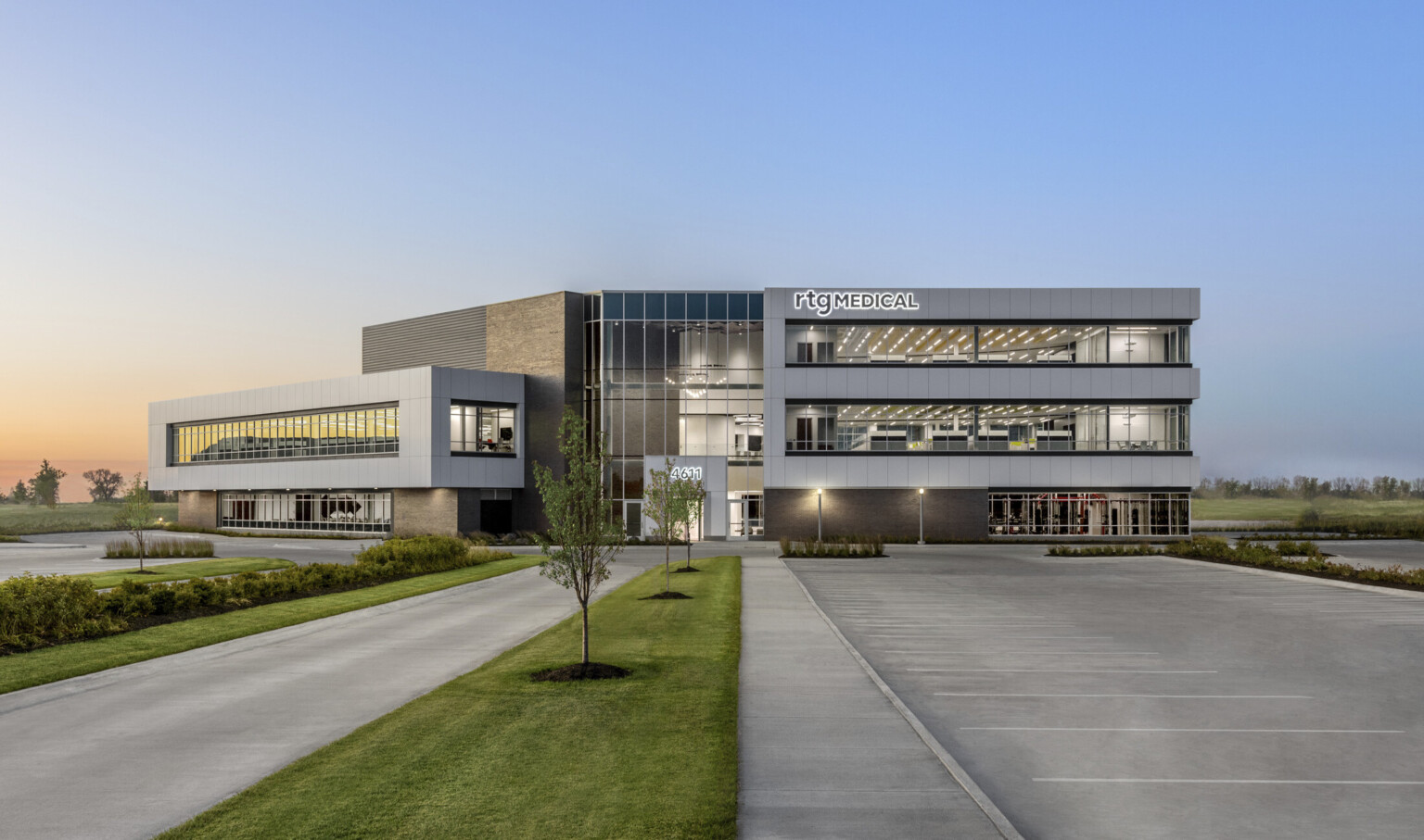 Front side of 3 story building illuminated in the early morning. Green space surrounding. Grey wrap facade accent, large windows