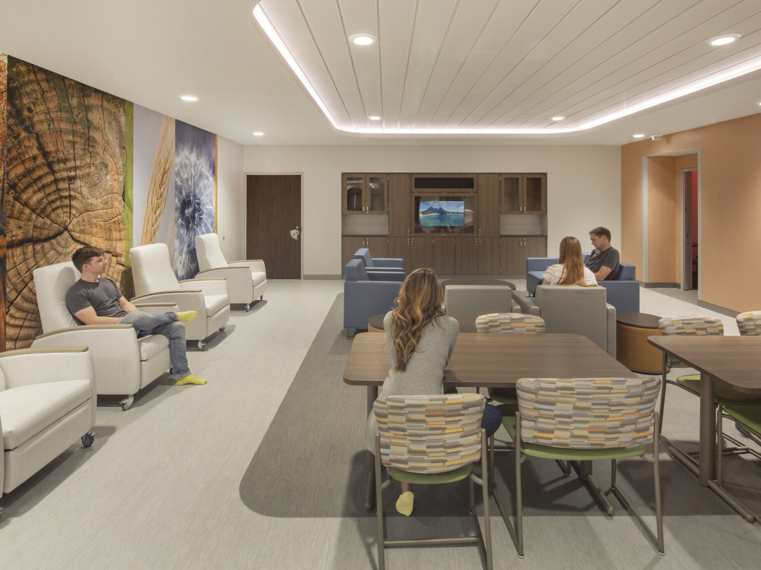 People in recliners and arm chairs seated in groups around tables in big room with illuminated recessed ceiling detail, orange wall, media screen