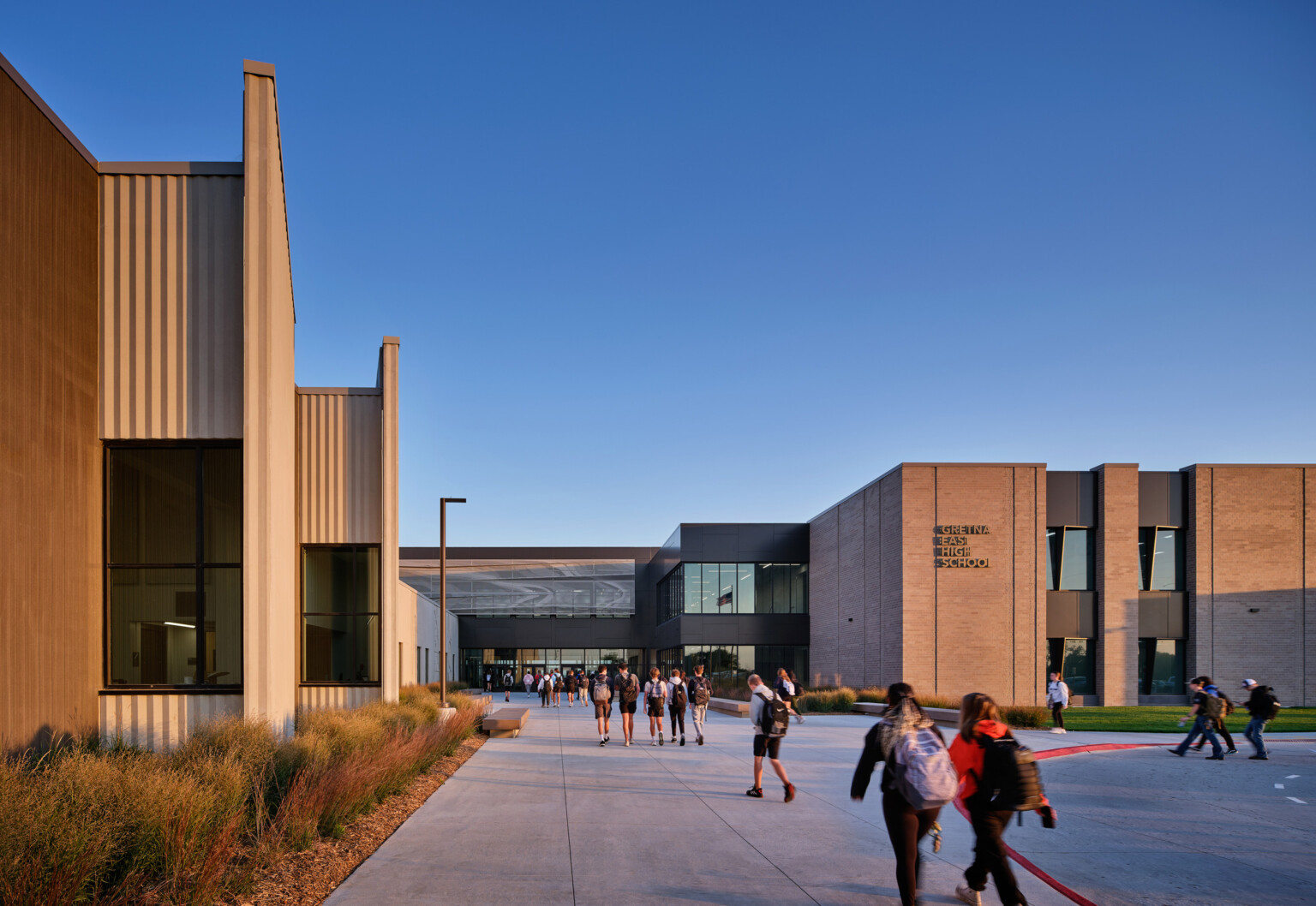 Multistory tan brick high school entry with large glass windows and students walking on walkways