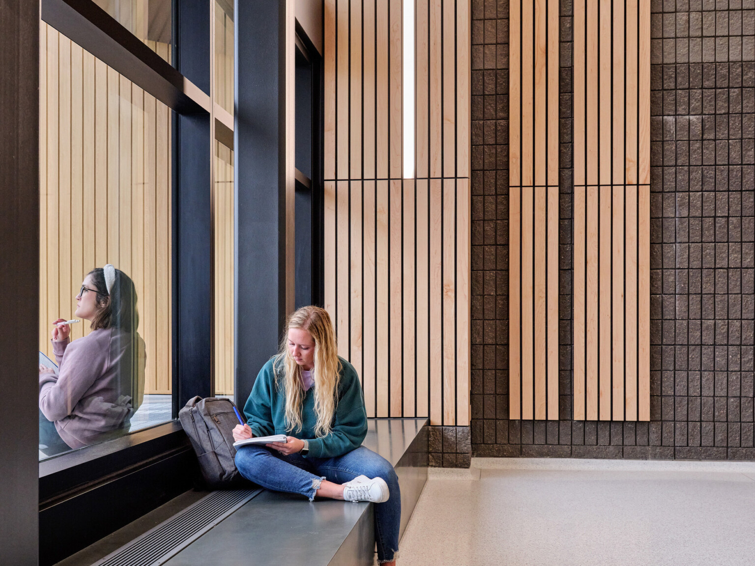 Girl in a green sweater sitting on a bench in front of a large window with a another girl in a purple sweater sitting on the outside of the window