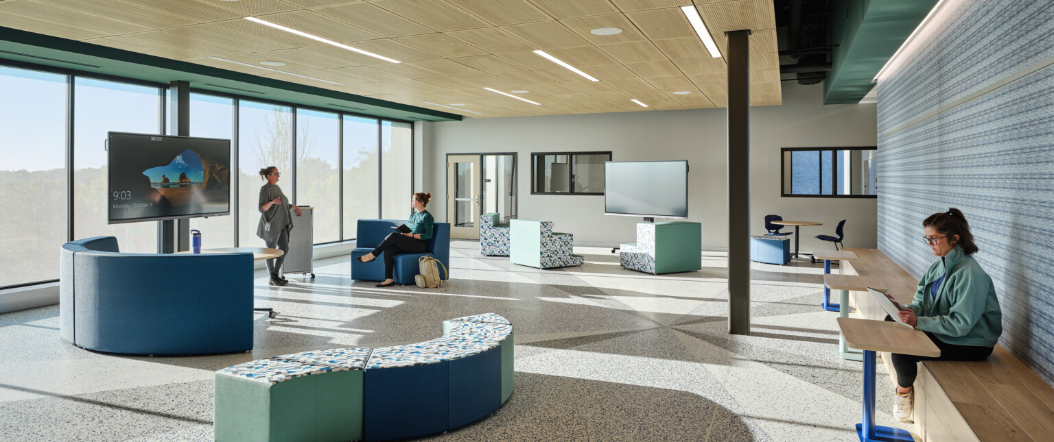 A gathering area on a school campus with modern blue and green seating with light wood ceiling, wall of windows, and a colorful accent wall