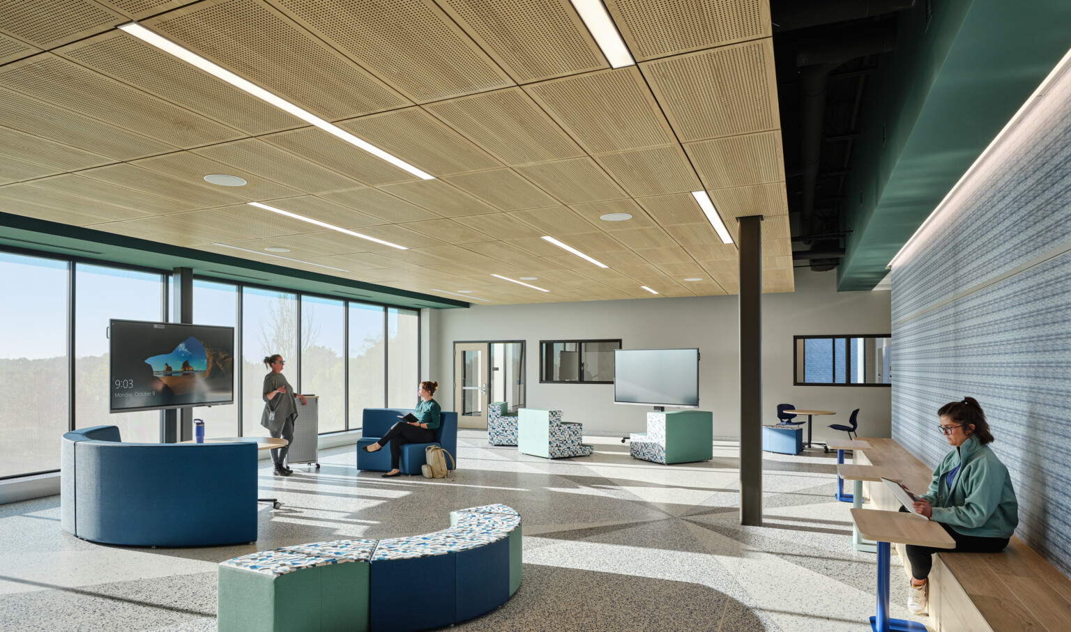 A gathering area on a school campus with modern blue and green seating with light wood ceiling, wall of windows, and a colorful accent wall