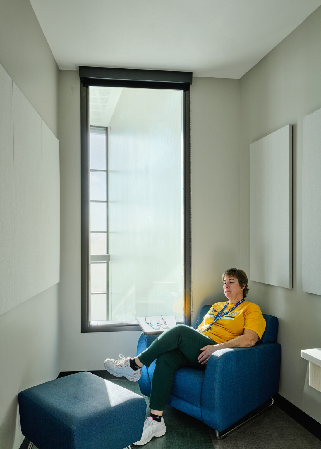 relaxation room in Boys Town Education Center; woman in a yellow shirt sitting in the corner comfy chair in a small closet like room with a large window behind her; room for trauma informed design relaxation room in Boys Town Education Center; woman in a yellow shirt sitting in the corner comfy chair in a small closet like room with a large window behind her; room for trauma informed design