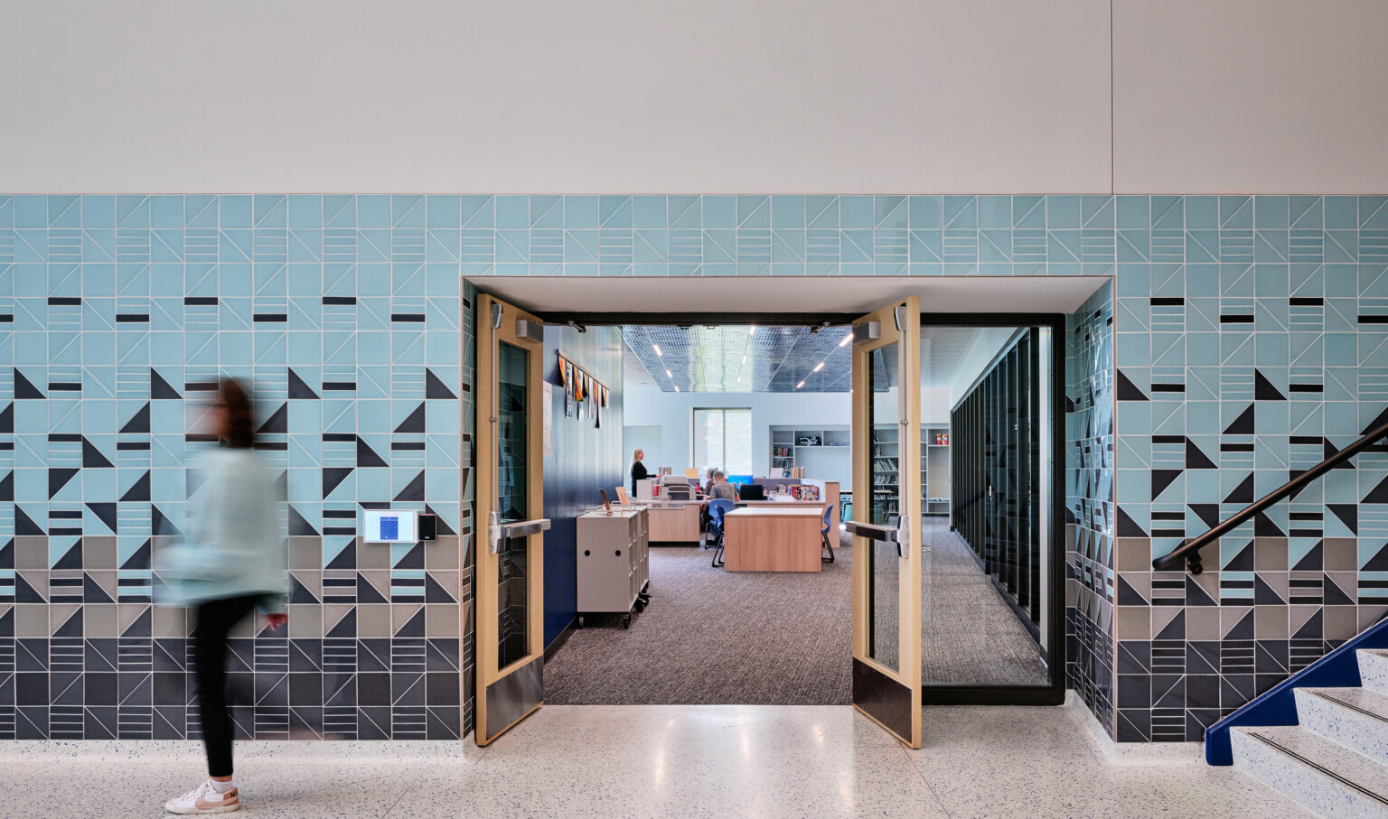 Abstract blue and grey tile wall surrounding a door opening looking into a office with light wood furniture