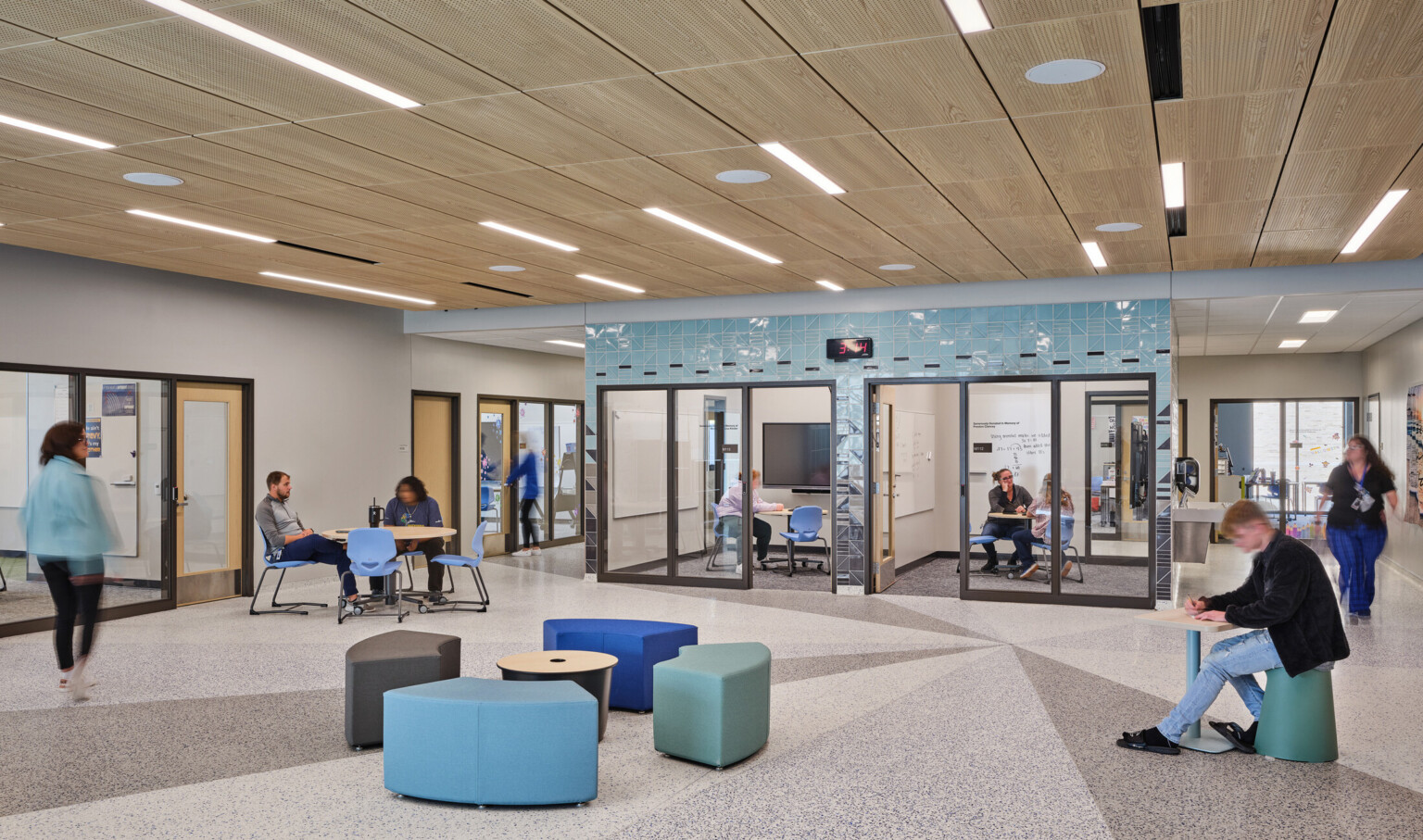 Open concept gathering area with blue and green modern seating filled with students surrounded by conference rooms with glass doors Open concept gathering area with blue and green modern seating filled with students surrounded by conference rooms with glass doors