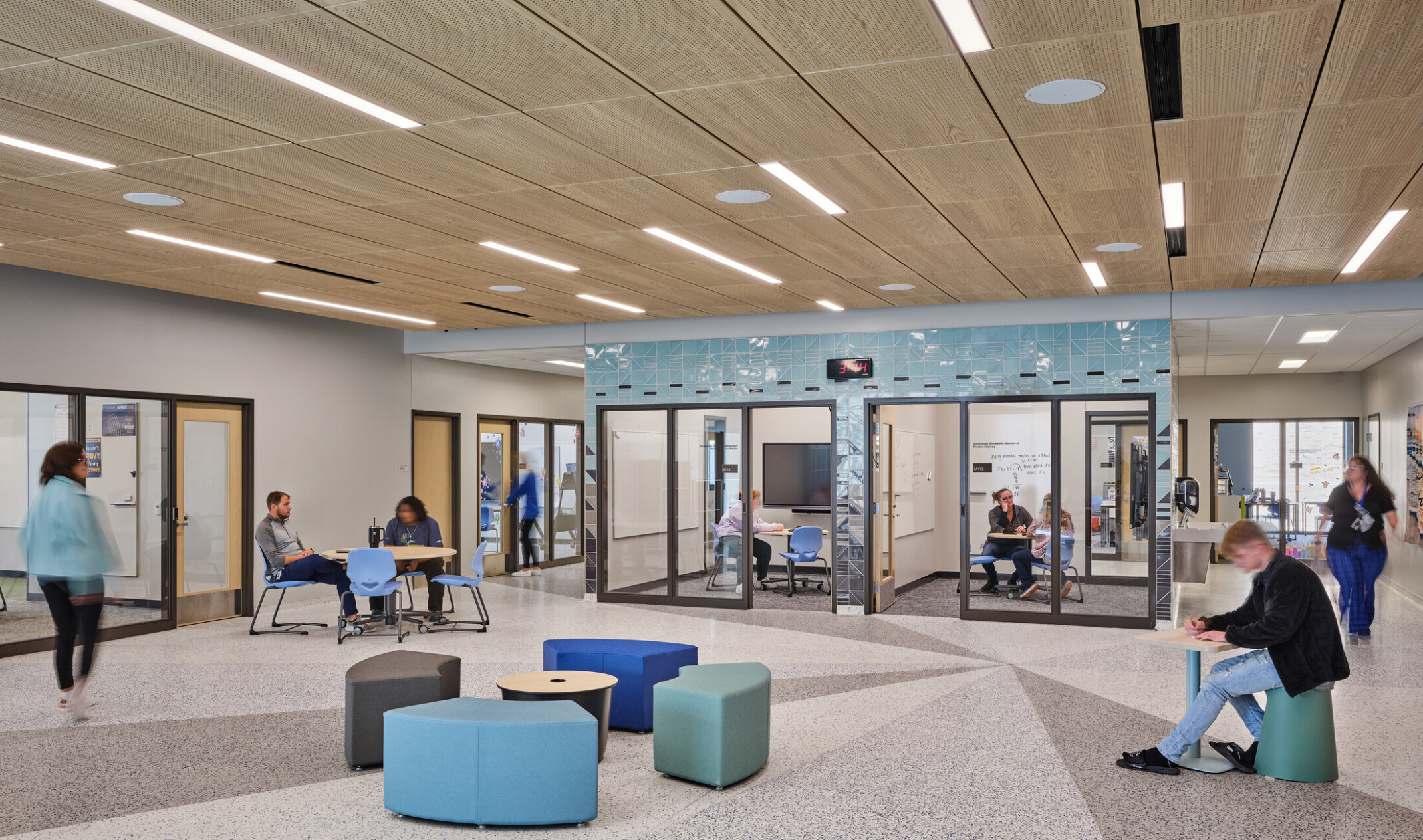 Open concept gathering area with blue and green modern seating filled with students surrounded by conference rooms with glass doors