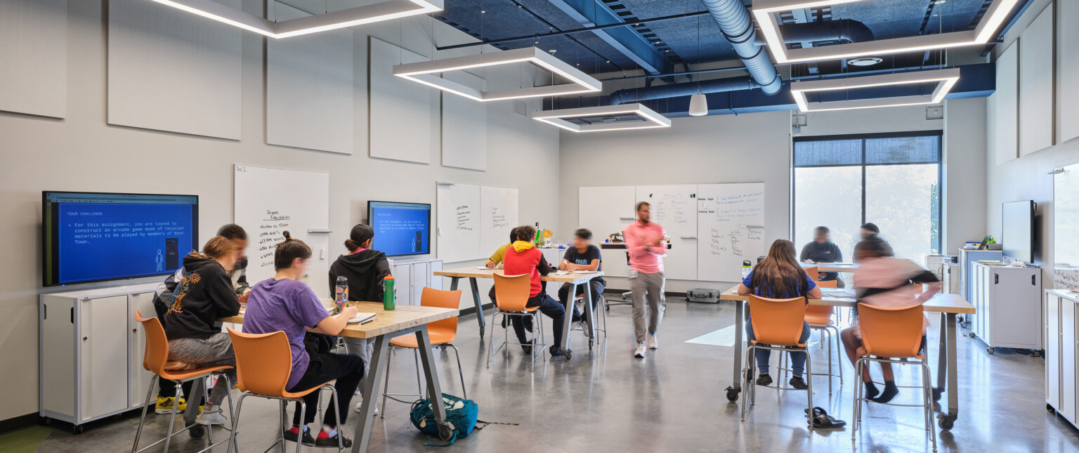 School classroom filled with students sitting at high tables with orange chairs. Modern square lighting is suspended from the ceiling