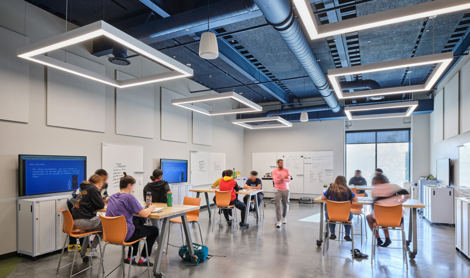 School classroom filled with students sitting at high tables with orange chairs. Modern square lighting is suspended from the ceiling