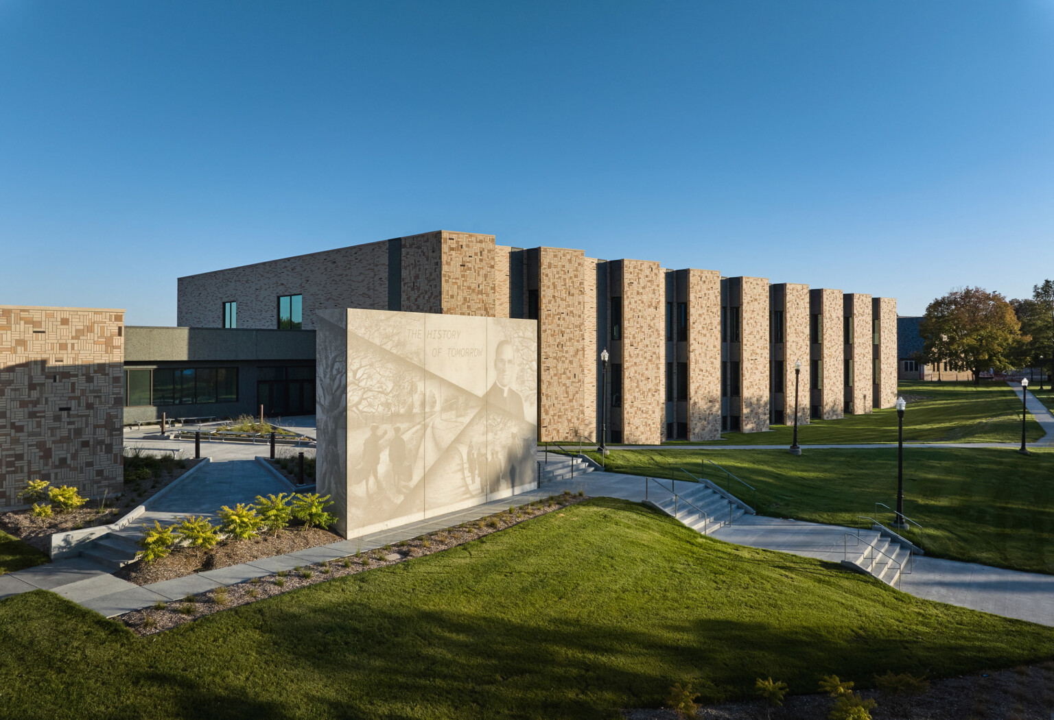 Boys Town Education Center exterior wall; red-tan stone with slated windows; shaded green space and walk paths in front of the wall; Stone structure with a mural light with phrase the history of tomorrow Boys Town Education Center exterior wall; red-tan stone with slated windows; shaded green space and walk paths in front of the wall; Stone structure with a mural light with phrase the history of tomorrow
