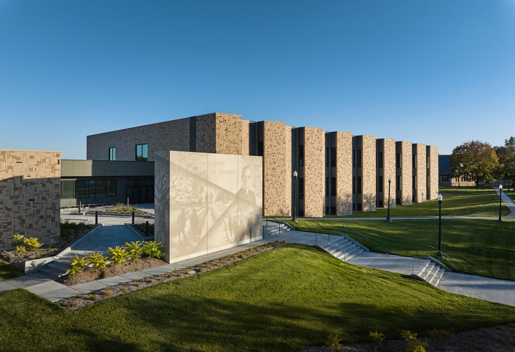 Boys Town Education Center exterior wall; red-tan stone with slated windows; shaded green space and walk paths in front of the wall; Stone structure with a mural light with phrase the history of tomorrow