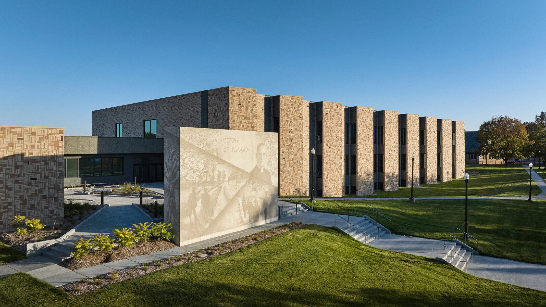 Boys Town Education Center exterior wall; red-tan stone with slated windows; shaded green space and walk paths in front of the wall; Stone structure with a mural light with phrase the history of tomorrow