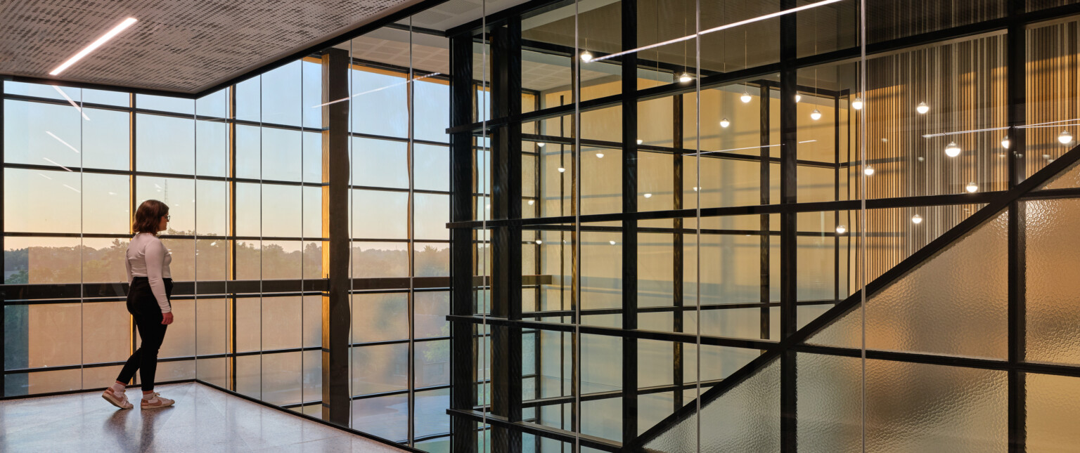 Girl in a white shirt and black pants standing in the corner of a room surrounded by windows