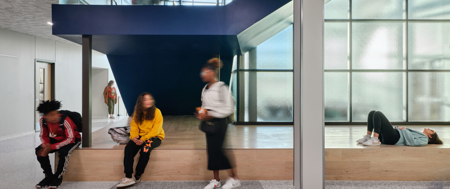 Kids gathered in a school common area under a blue staircase with a light wood raised seating area