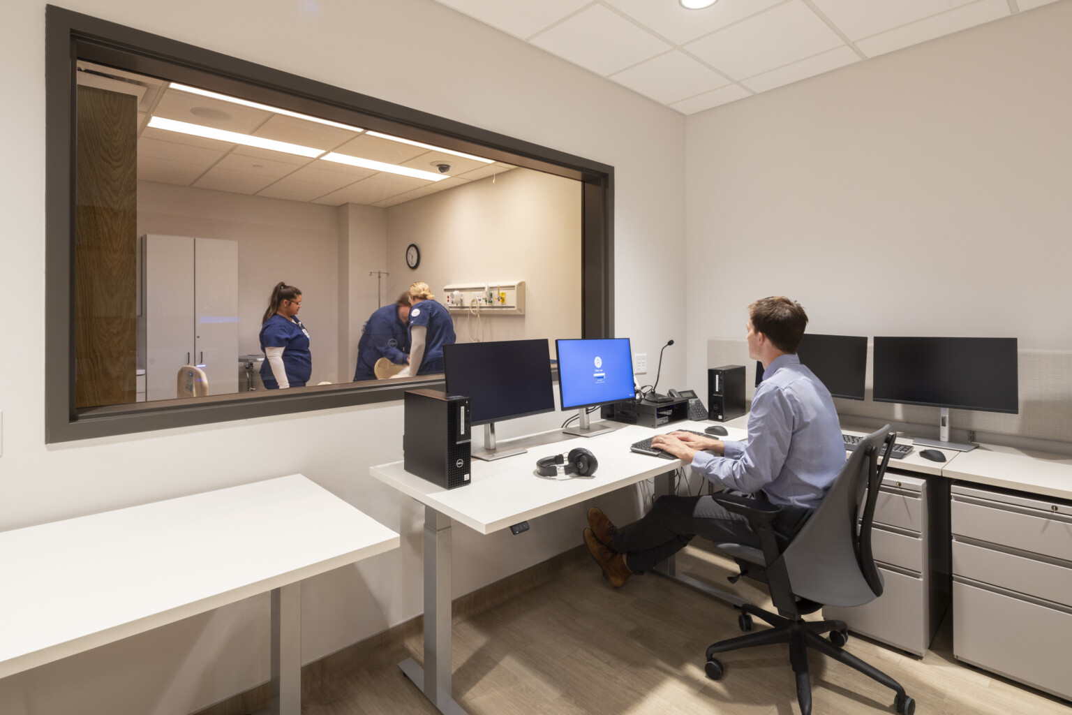students in a technical learning space; one student behind glass window on computers facing medical professionals on the other side of glass