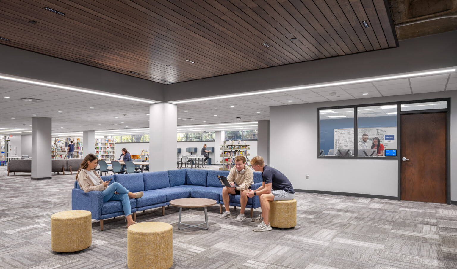 modern library lounge area; gray patterned carpet and a wood-paneled ceiling; blue sectional sofa; three round ottomans arranged around a small circular table in the foreground; few people are seated and reading; background bookshelves and study tables; a glass windowed office door on the right; brightly lit with recessed ceiling lights