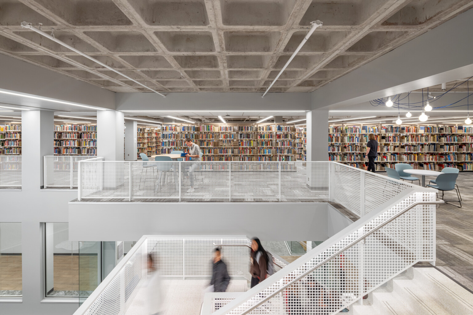 spacious, modern library interior; an open layout, multiple levels; upper-level features rows of bookshelves with books along the back wall; tables and chairs for reading and studying; foreground shows a white metal staircase, railing leading to the lower level; people walking; ceiling exposed concrete grid design; space brightly lit with overhead lights.
