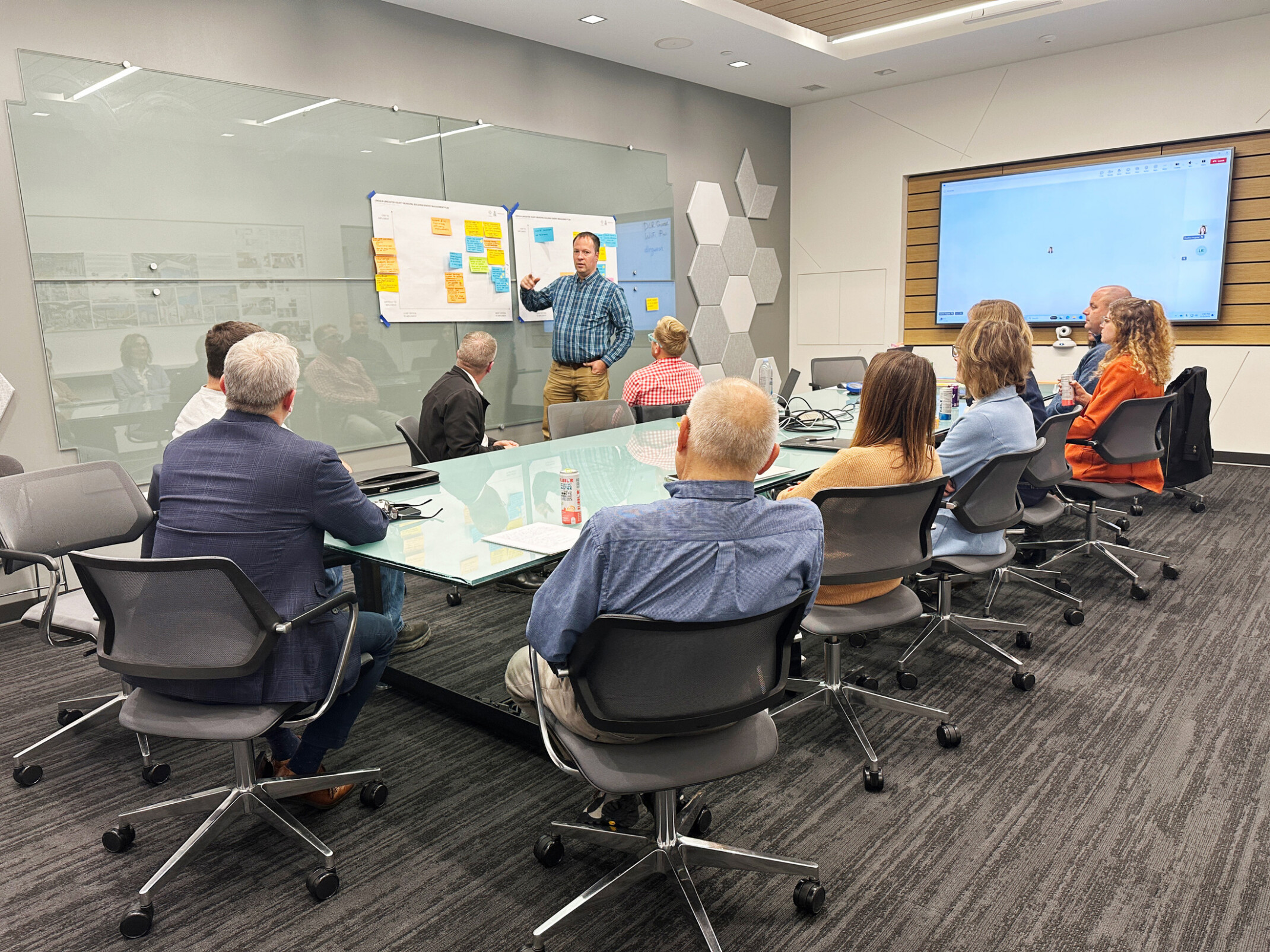 A group of professionals sit at a conference table looking at a man standing in front of posterboards with sticky notes on them.