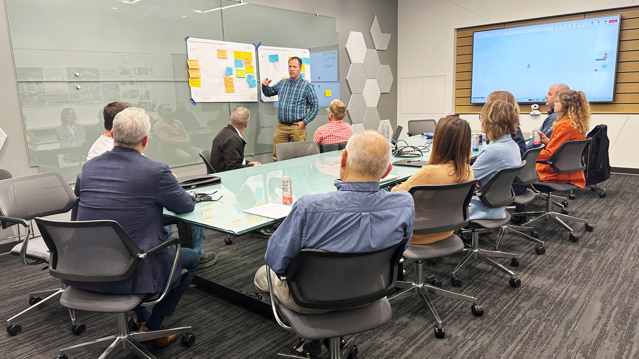 A group of professionals sit at a conference table looking at a man standing in front of posterboards with sticky notes on them.