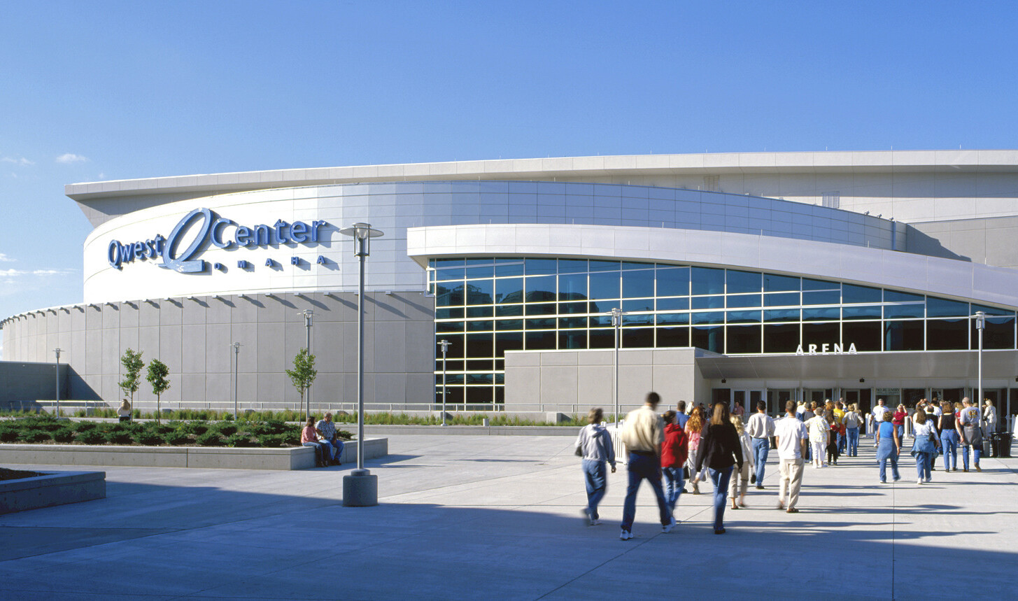 Glass facade entry with curved white roof in front of a round building section with metal panels, labeled Qwest Center Arena