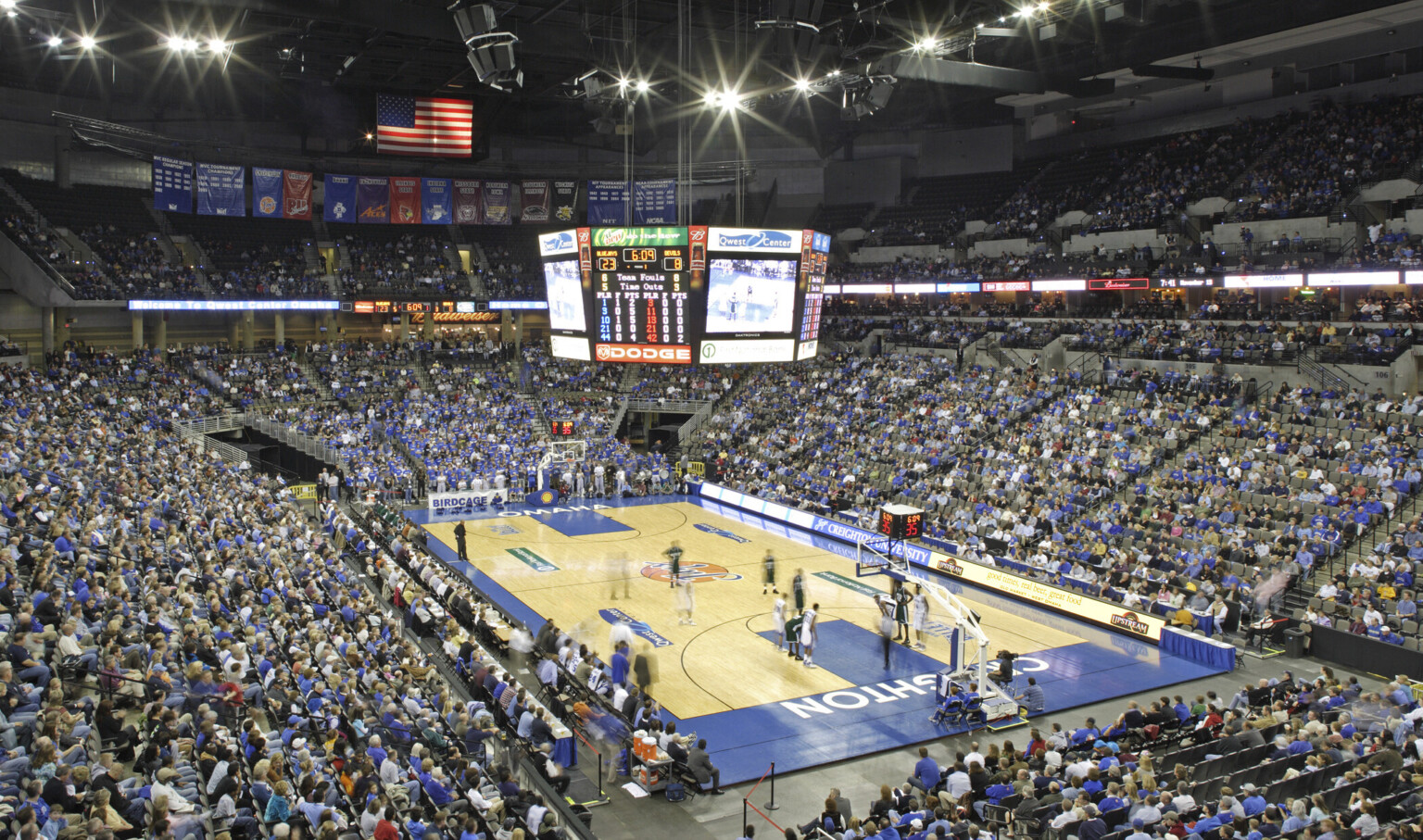 Basketball court with lighting and jumbotron hanging above. Bleachers surround the court and are filled with fans