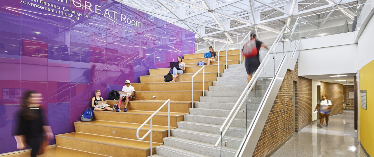 main thoroughfare indoor stairway with adjacent bleachers for seating, white steel girders support triple-height glass ceiling and skylights, day lit grand room