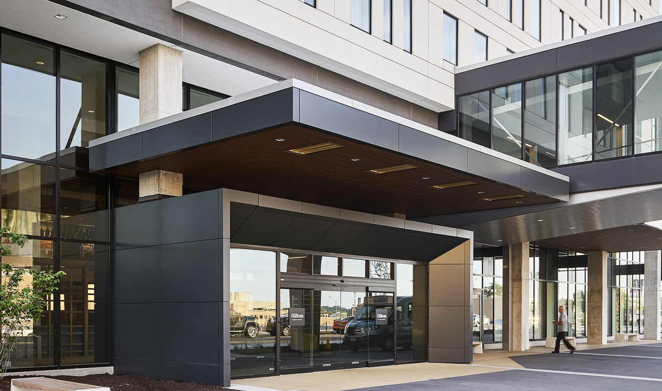 Entryway with benches out front, an overhang, and glass doors wrapped in dark framing at hilton des moines
