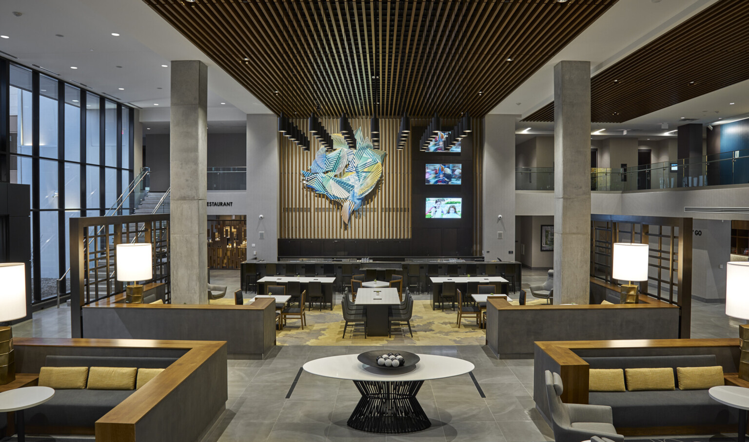 Mezzanine hallway in atrium with bar and tables at front of lower level. Stone pillars center and side 2nd floor walkways